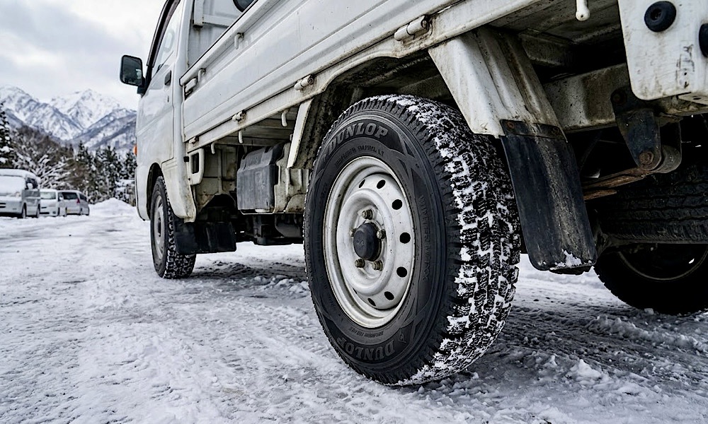 雪の降る早朝の物流拠点に停まる小型トラックとスタッドレスタイヤ